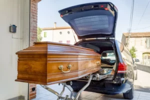 Un cercueil en bois verni en cours de chargement à l'arrière d'un corbillard noir.